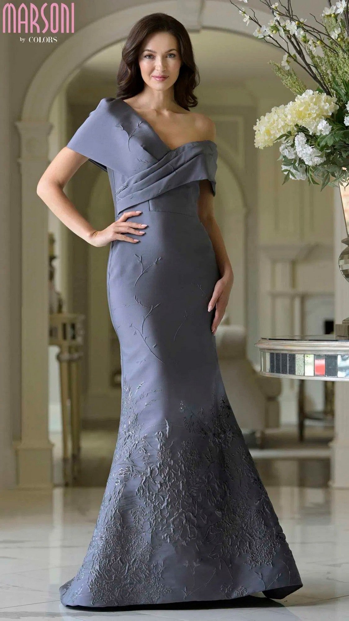 A woman poses indoors with one hand on her hip, wearing the Long Formal Dress: Marsoni MV1378 in charcoal—a fitted, asymmetric off-shoulder evening gown with textured floral details.